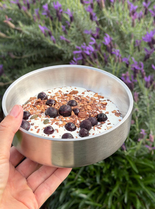 A stainless steel bowl filled with yogurt or cream, topped with chocolate pieces and chocolate crumbs, held by a hand against a blurred background of green grass and purple flowers.