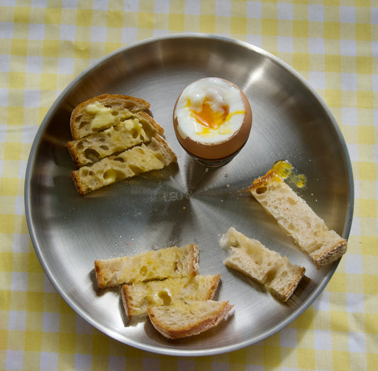 A stainless steel plate on a yellow and white checkered tablecloth, holding several slices of buttered bread and a hard-boiled egg with a runny yolk.