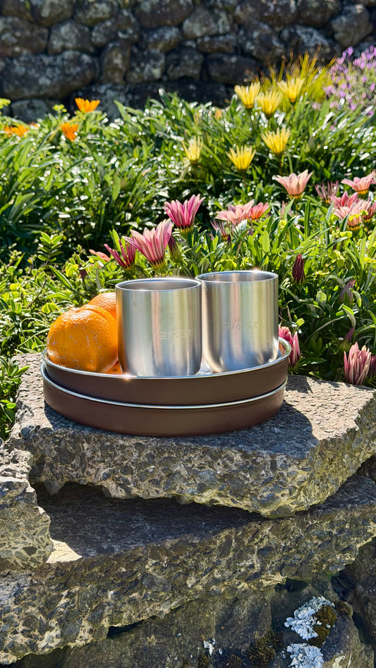 Two stainless steel travel mugs sitting on a brown ceramic tray, placed on a stone ledge outdoors with flowers and a stone wall in the background.