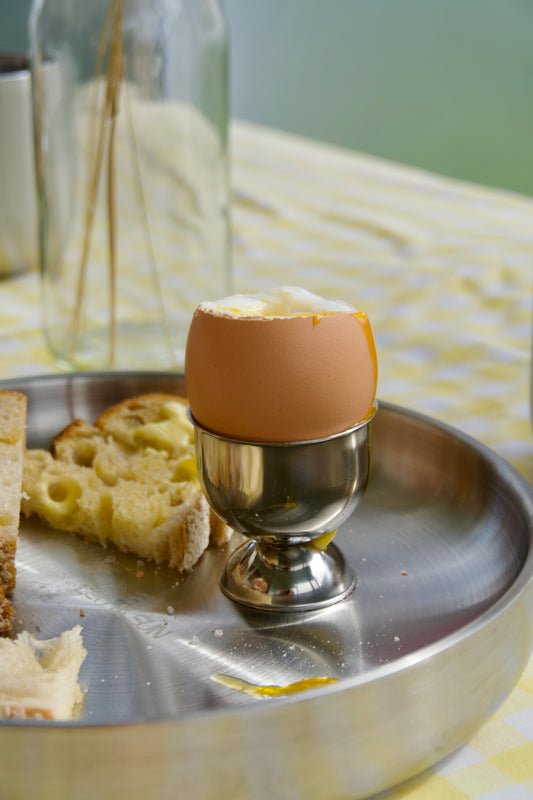 A cooked egg half-shell with butter sits in a small metal egg cup on a round metal tray, alongside sliced bread on a yellow-and-white checkered tablecloth.