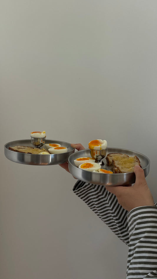 Two stainless steel plates held by a person wearing a black and white striped shirt, containing a breakfast spread with eggs, toast, and a small drink, against a plain light gray wall.