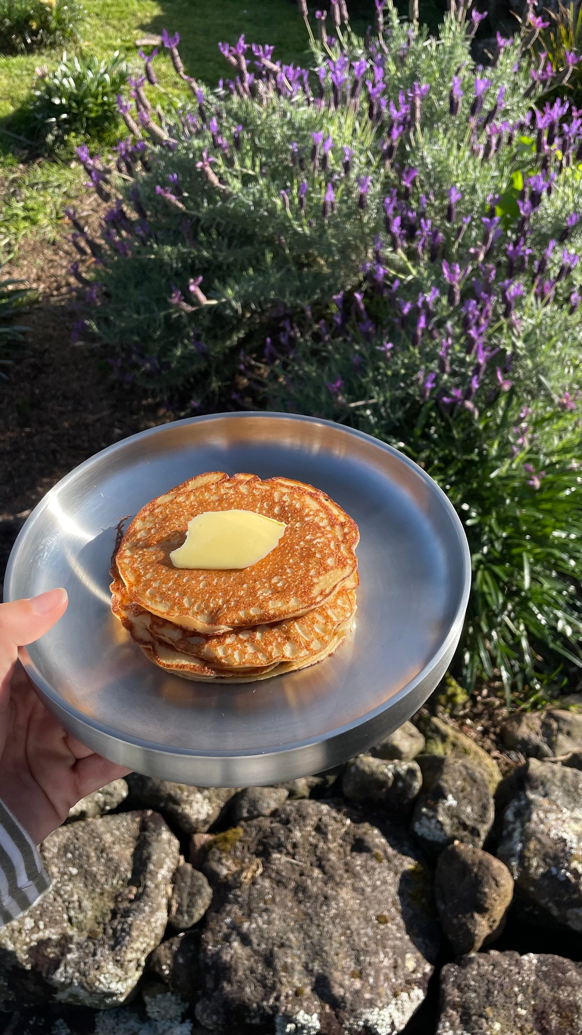 A stack of golden-brown pancakes topped with a pat of butter on a silver plate, held by a hand outdoors in front of purple flowers and rocks.