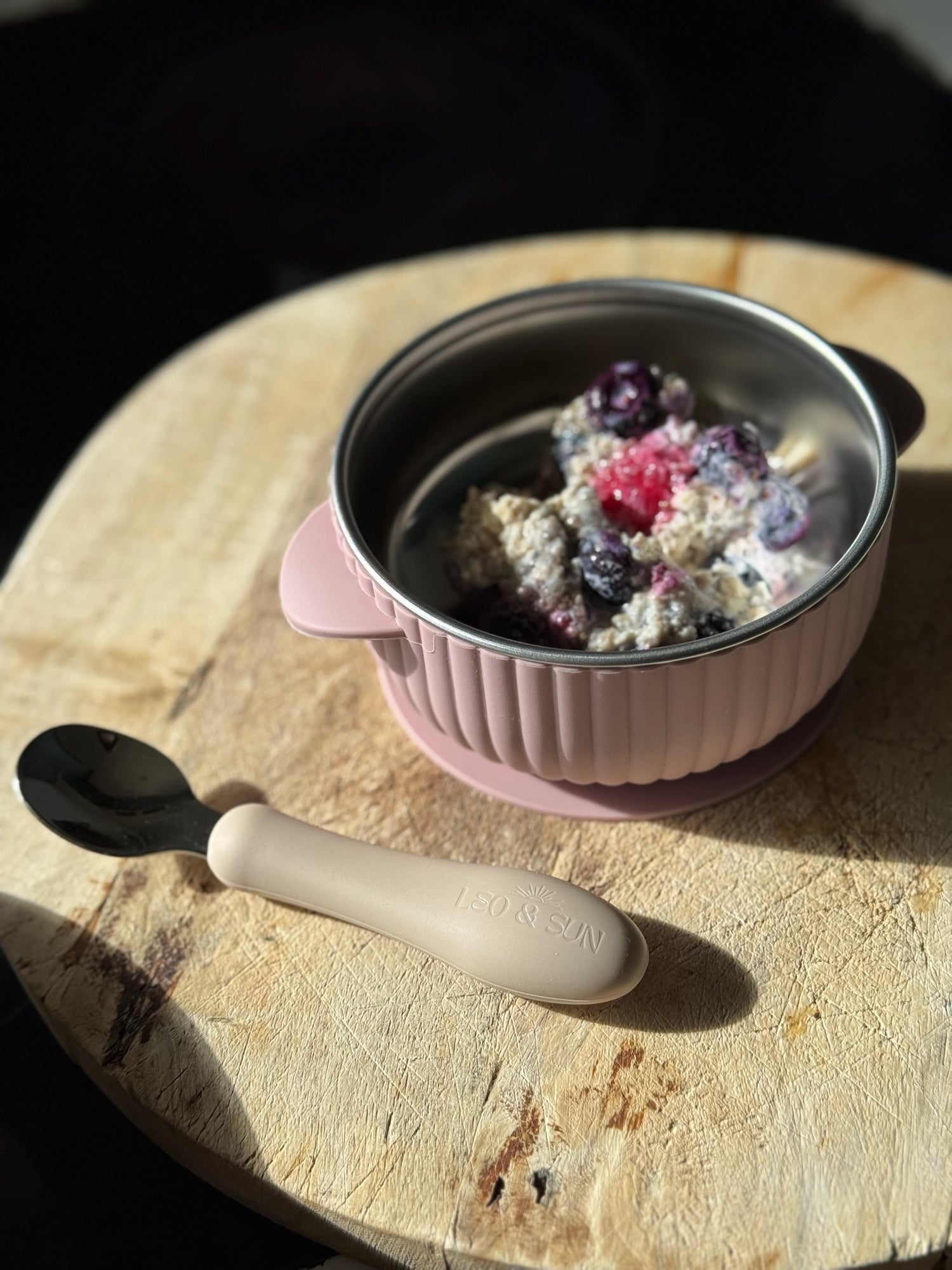 Pink baby bowl with silver lid containing a dessert on a wooden surface, accompanied by a beige spoon.